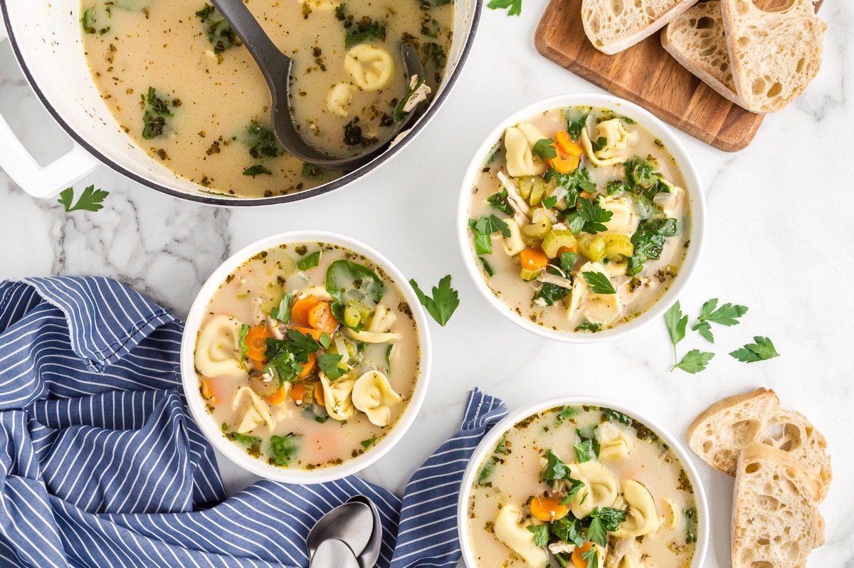 Several bowls of soup with cheese tortellini, shredded chicken, carrots, celery, spinach, and parsley arranged on a white surface with bread nearby.