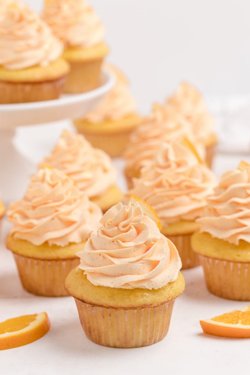 A close-up of freshly frosted Dreamy Orange Cupcakes with Whipped Orange Frosting, with more cupcakes displayed on a cake stand in the background.