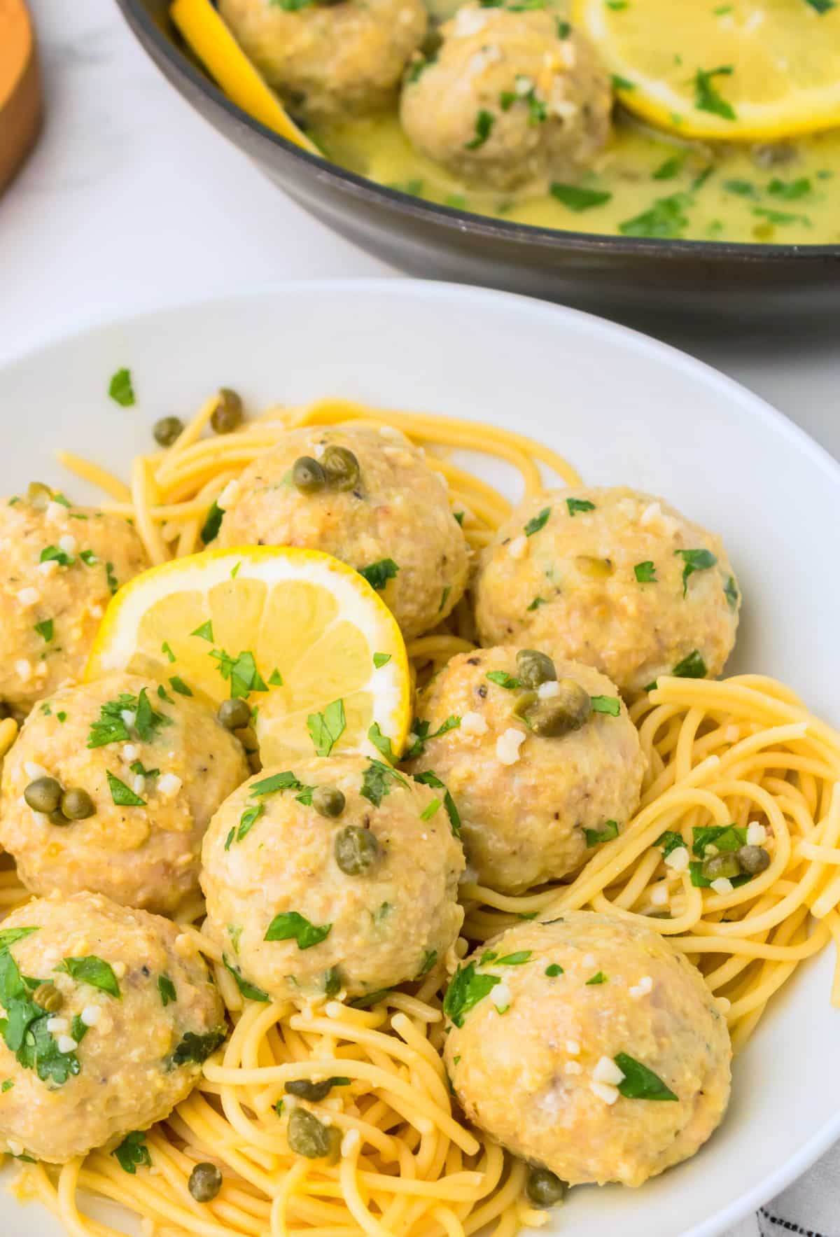  Plate of chicken piccata meatballs over pasta with skillet of extra meatballs in the background