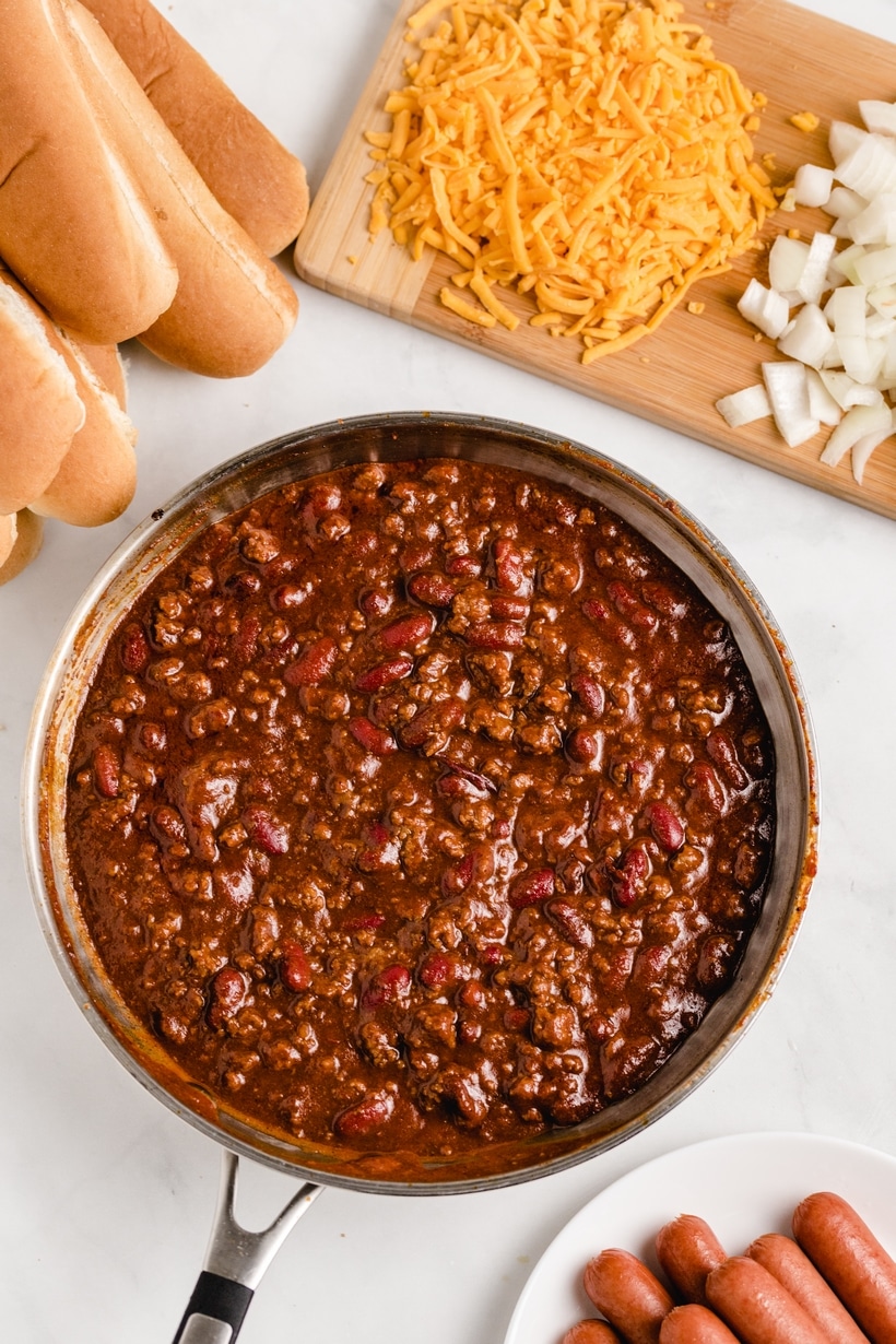 Overhead shot of hot dog chili in a pot with buns, shredded cheese, and onions on the side 