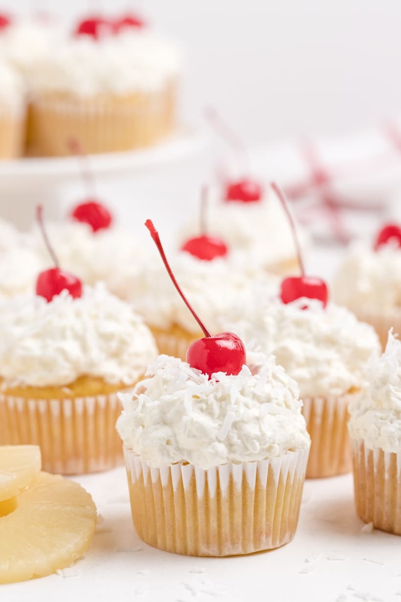 A batch of Piña Colada Cupcakes topped with coconut whipped cream, shredded coconut, and maraschino cherries, arranged on a serving tray.