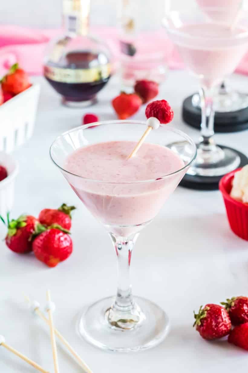 Two martini glasses filled with the berries and cream frozen martini, placed on a table with fresh berries scattered around.