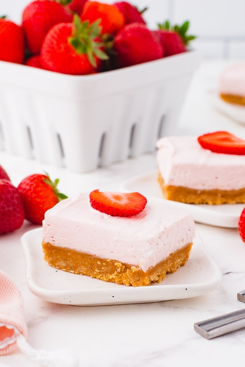 Two no-bake strawberry cheesecake bars on white plates with fresh strawberries in the background