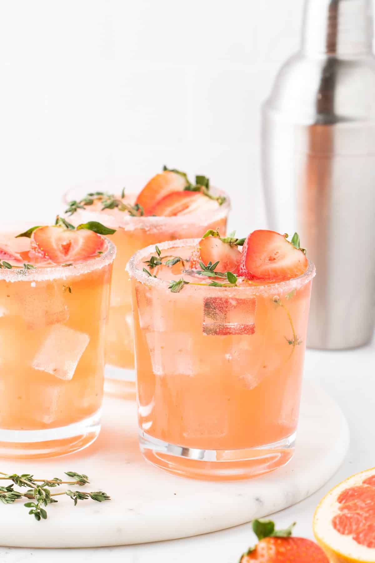 Strawberry Grapefruit Paloma on a marble tray Three palomas on a white marble round with a cocktail shaker and citrus slices in the background.
