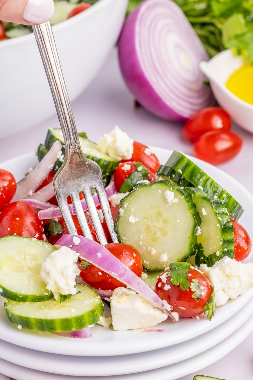 Cucumber Salad with Feta and Red Onion Served Cold Plate of cucumber tomato onion salad with feta and a fork, shown with lemon and fresh herbs in the background.