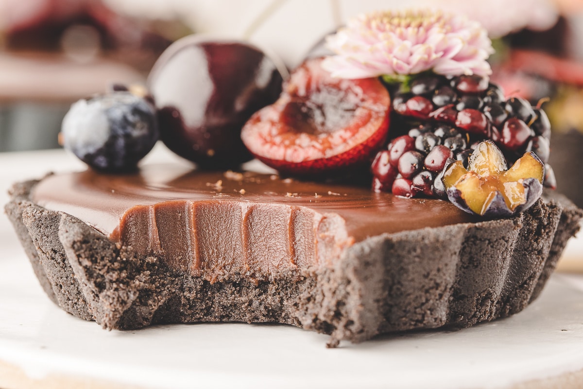 Side angle of a mini chocolate ganache tart with a clean bite taken out, showing the smooth ganache and cookie crust layers.