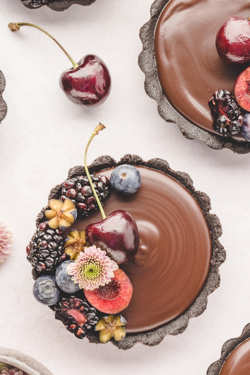 Top-down view of a mini chocolate ganache tart topped with fresh berries and edible flowers on a white background.