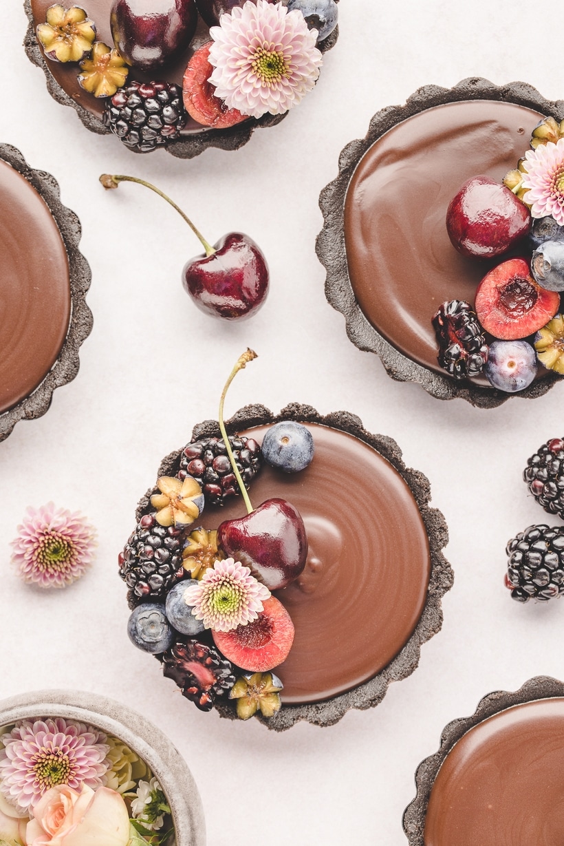 Top-down photo of multiple mini chocolate ganache tarts arranged on a surface, some decorated and some plain.