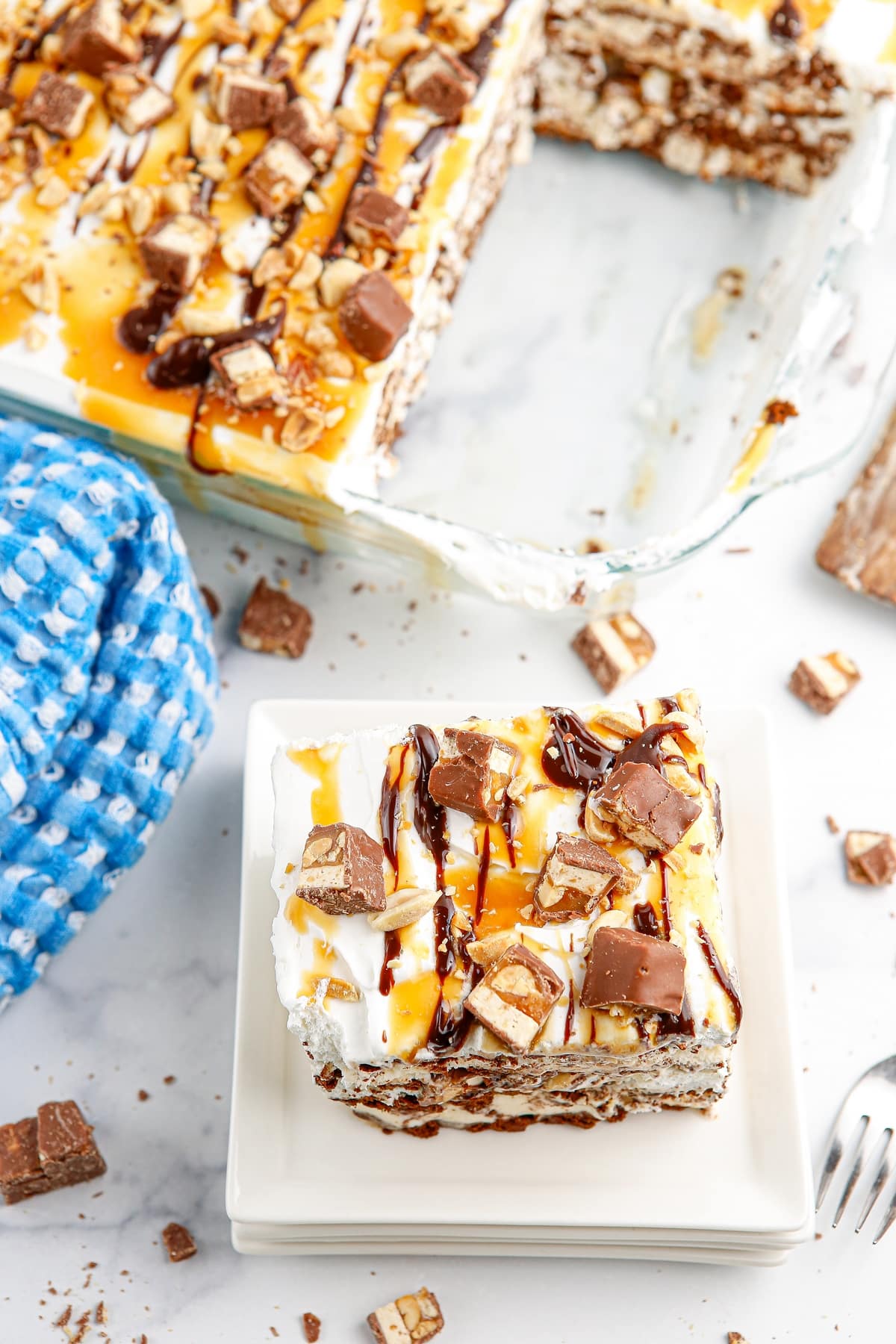 snickers ice cream casserole in a 9x13 pan with slice removed Snickers ice cream casserole topped with caramel, chocolate sauce, chopped peanuts, and Snickers bars