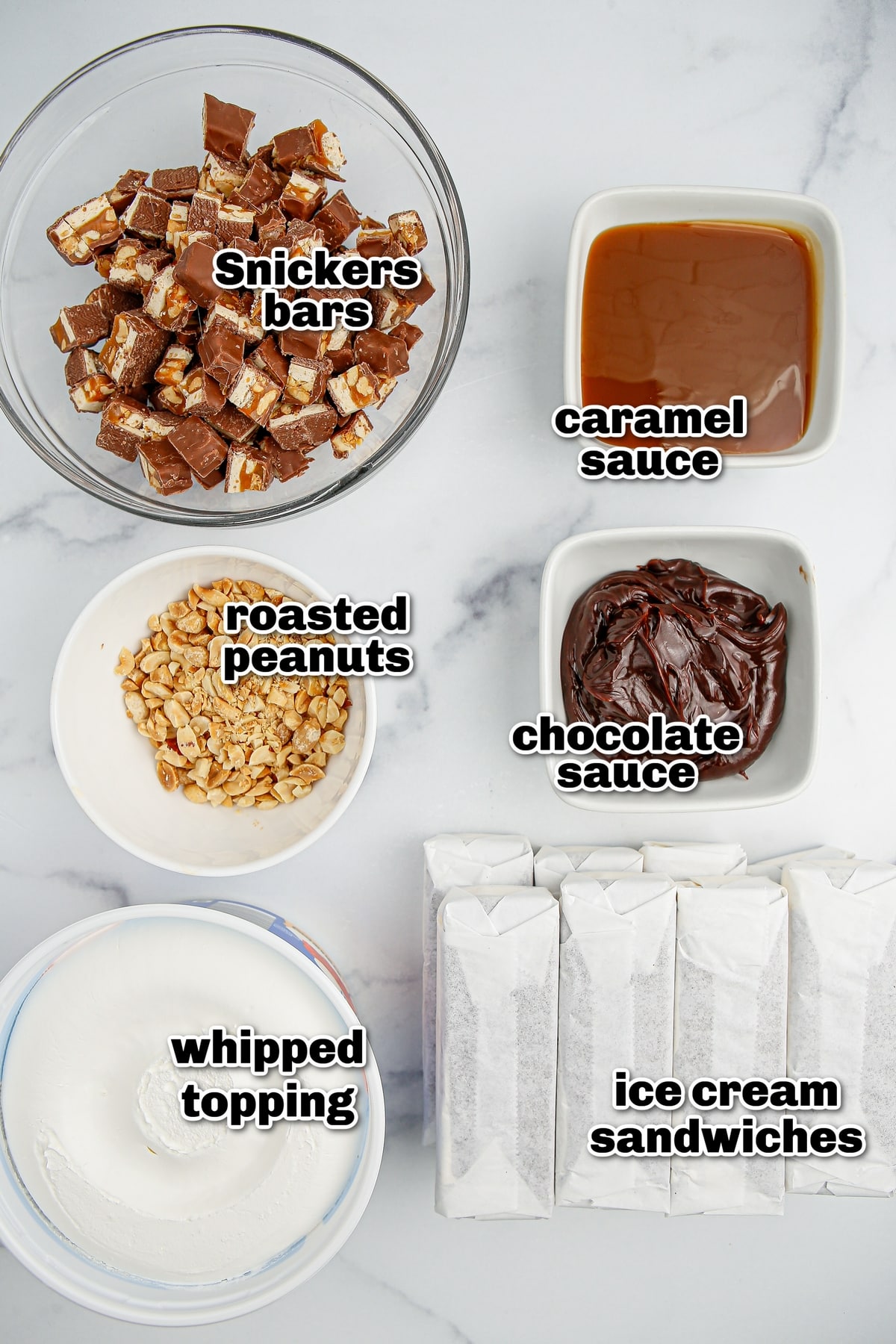 snickers ice cream sandwich cake ingredients laid out on counter Overhead view of ingredients for snickers ice cream cake including whipped topping, chocolate and caramel sauces, peanuts, Snickers bars, and ice cream sandwiches