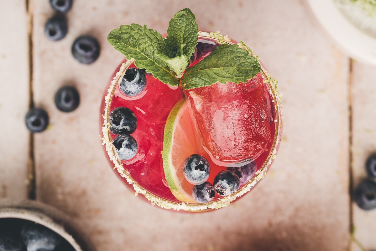 Overhead view of a blueberry margarita filled with ice, lime slices, and blueberries.