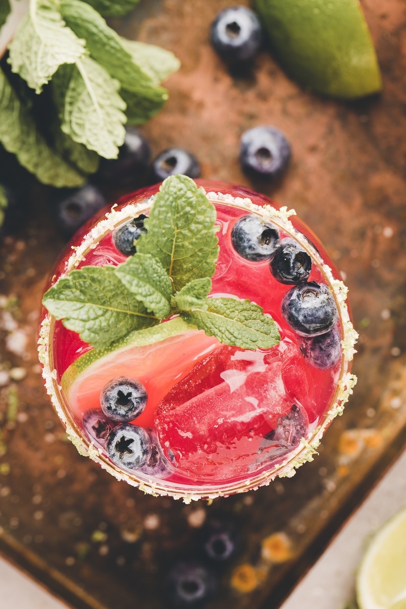 Blueberry margarita mix being poured from a shaker through a strainer into a glass filled with ice.