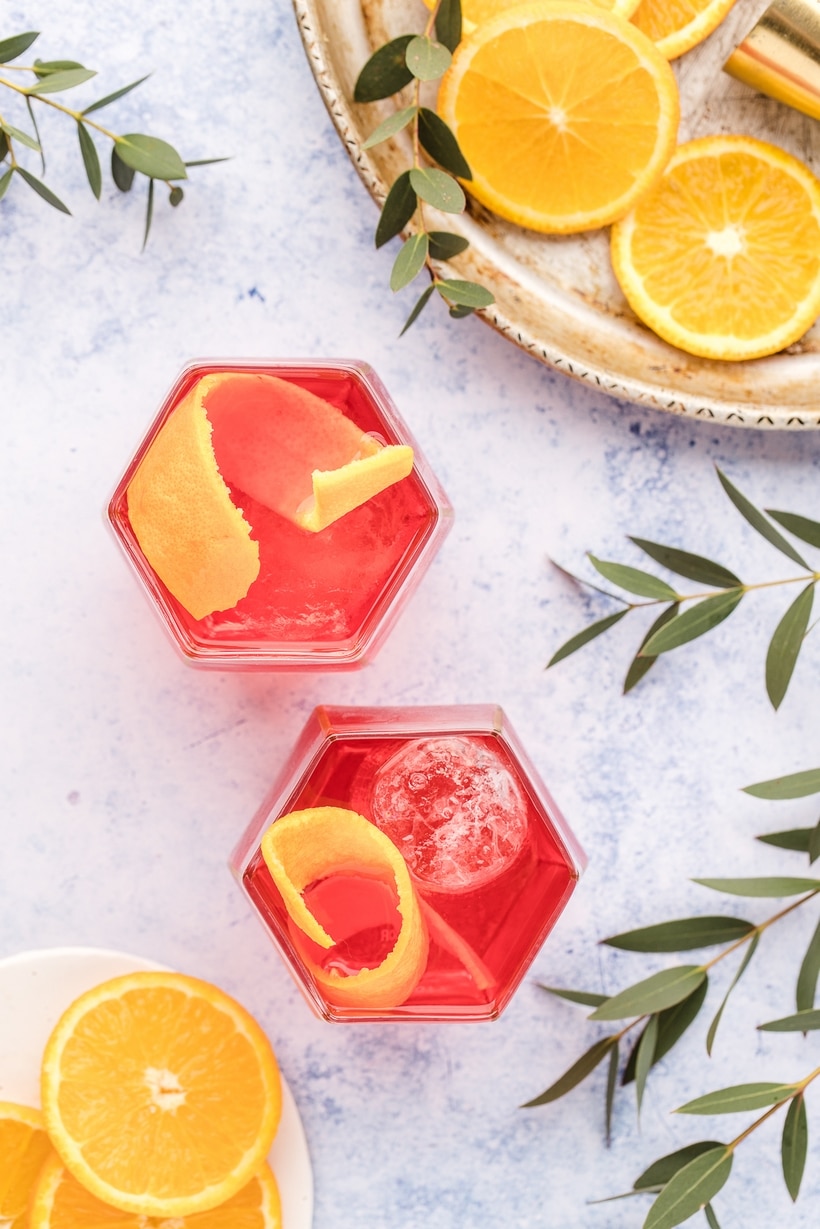 Negronis with orange peel and fresh orange slices Overhead view of gin-forward Negronis with orange peel garnish and sliced oranges on a tray.