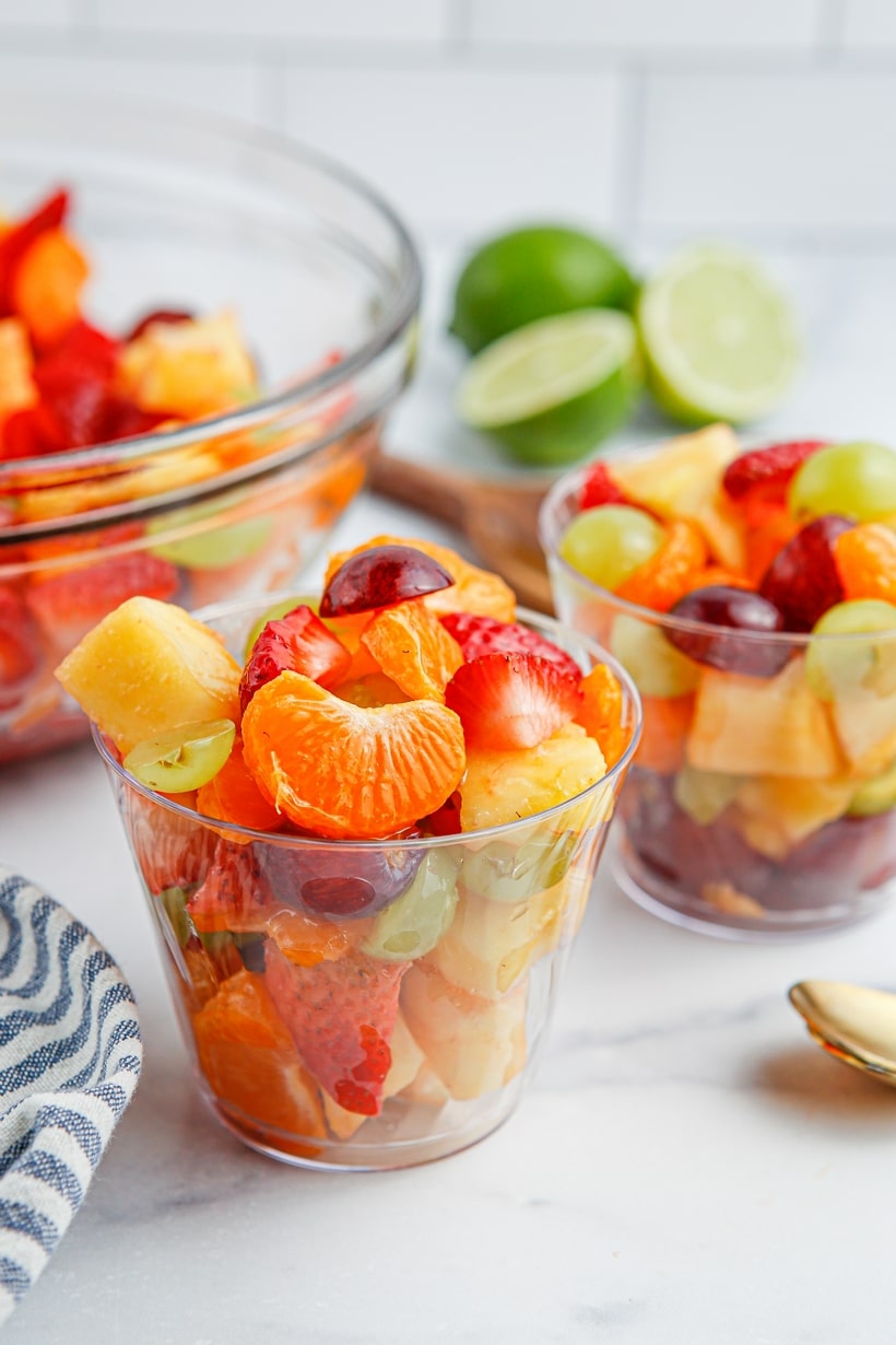 Honey Lime Fruit Salad in two plastic cups Honey lime fruit salad served in clear cups with whole and halved limes placed nearby, adding a pop of green to the colorful scene.