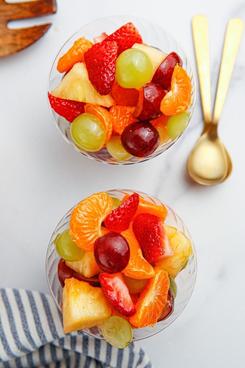 Honey Lime Fruit Salad with gold spoons on the side Overhead view of two fruit salad-filled cups on a surface, each with a gold spoon placed on top, ready for serving.