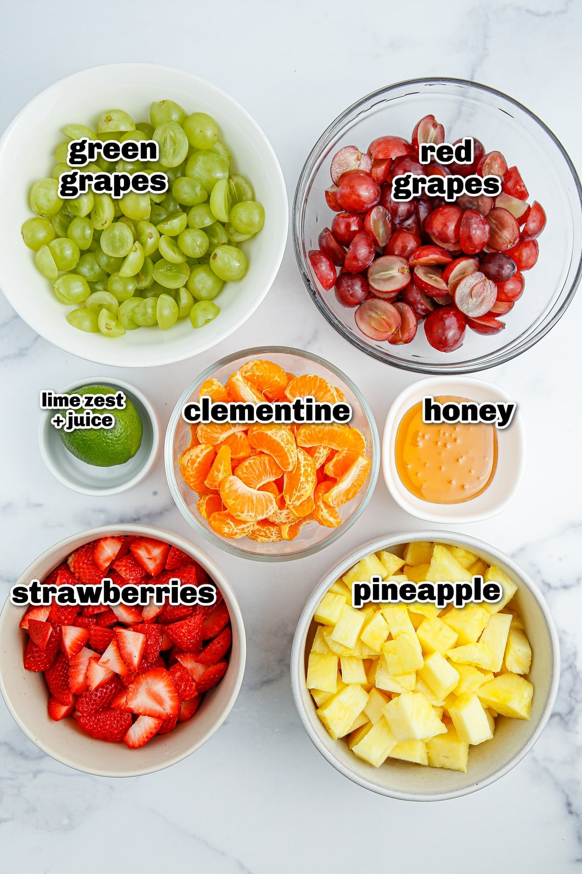 Honey Lime Fruit Salad ingredients All the fruit salad ingredients laid out on a surface, including grapes, pineapple, strawberries, clementines, a lime, and a small bowl of honey.