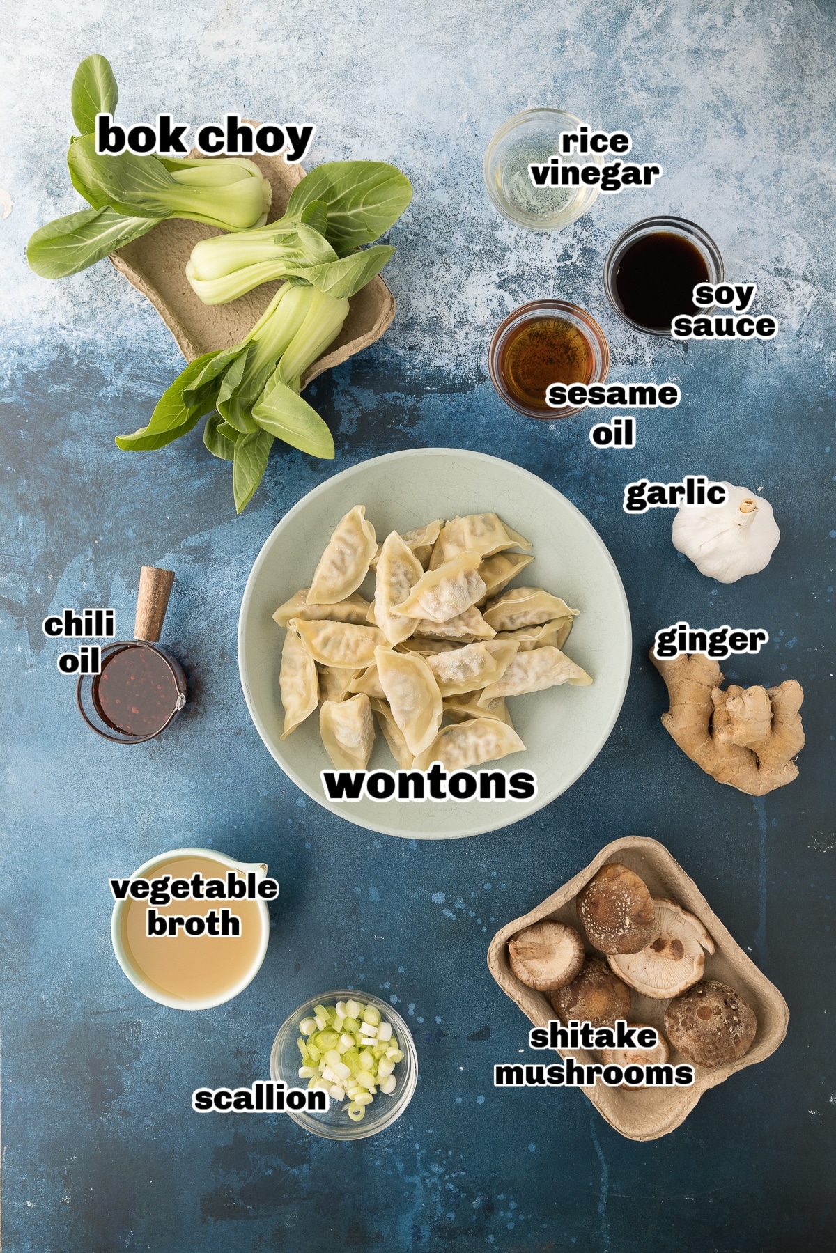 Ingredients for wonton soup arranged on a countertop, including sesame oil, bok choy, garlic, ginger, mushrooms, broth, frozen wontons, rice vinegar, soy sauce, scallions, sesame seeds, and chili oil.