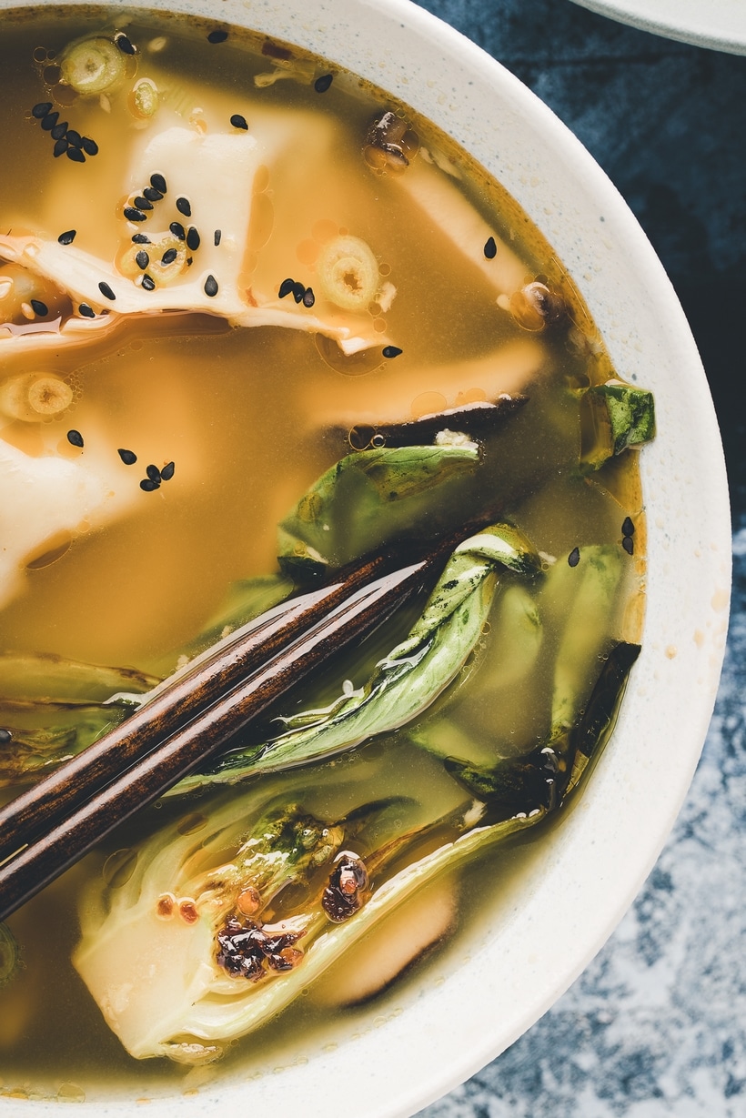 Close-up of a steaming bowl of wonton soup with tender wontons, bok choy, and garnishes of scallions, sesame seeds, and chili oil, served with chopsticks resting on the bowl.