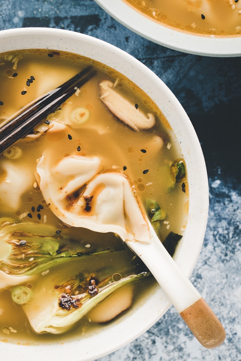 A bowl of wonton soup with tender wontons, bok choy, and a flavorful broth, garnished with scallions, sesame seeds, and chili oil.