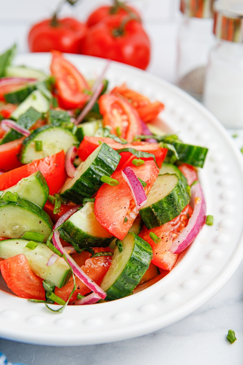 Fresh cucumber tomato salad with red onion and herbs in white dish, vine tomatoes in background