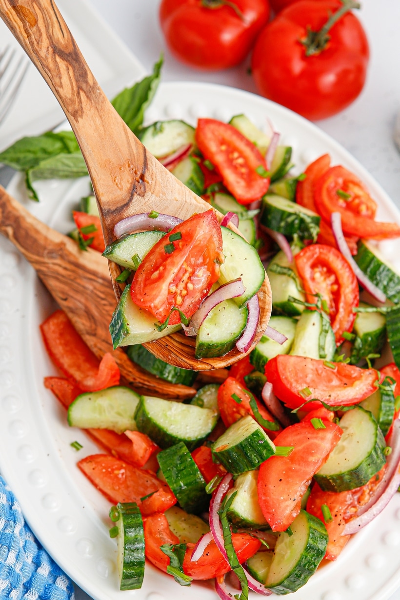 Close-up of a sliced tomato and cucumber on a wooden spoon with red onion and herbs