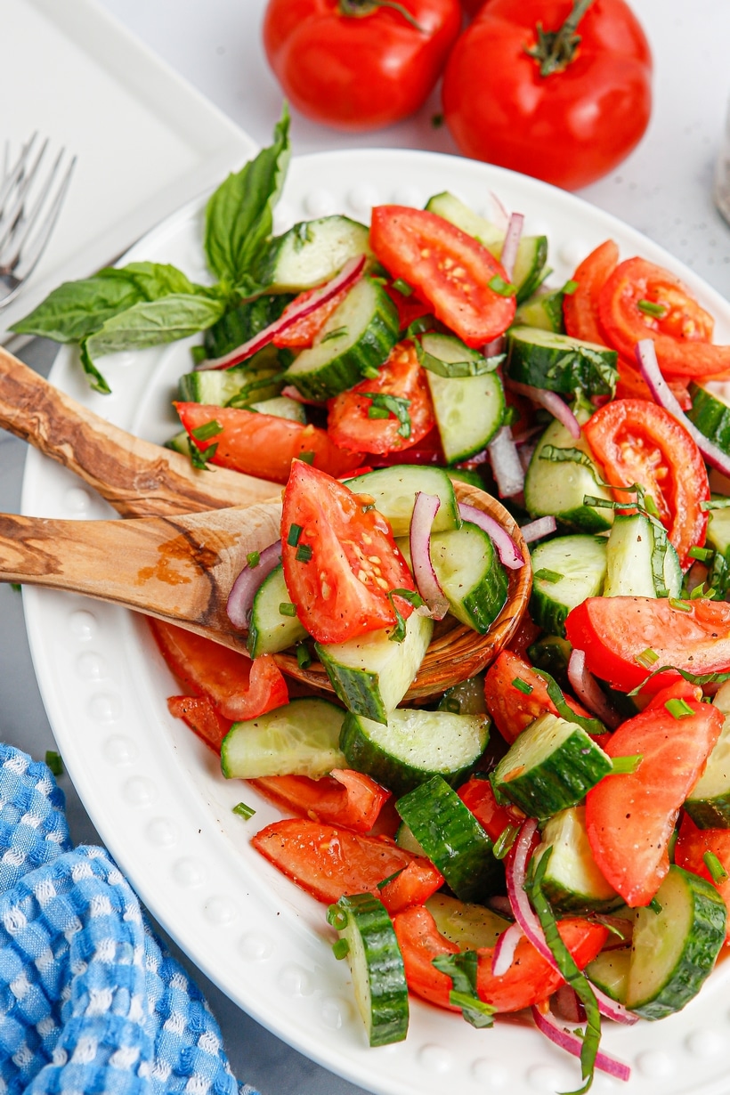Overhead angled shot of cucumber tomato salad in white dish with fresh basil and blue towel