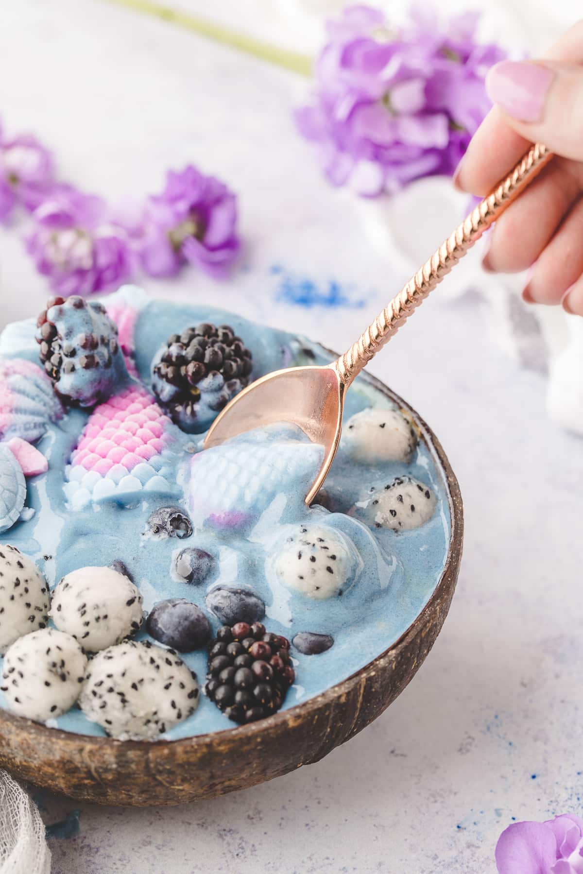 A spoonful of creamy smoothie with fresh fruit and yogurt mermaid decorations. Spoon scooping a bite of a mermaid smoothie bowl with yogurt mermaid tails, dragon fruit, blueberries, and blackberries.