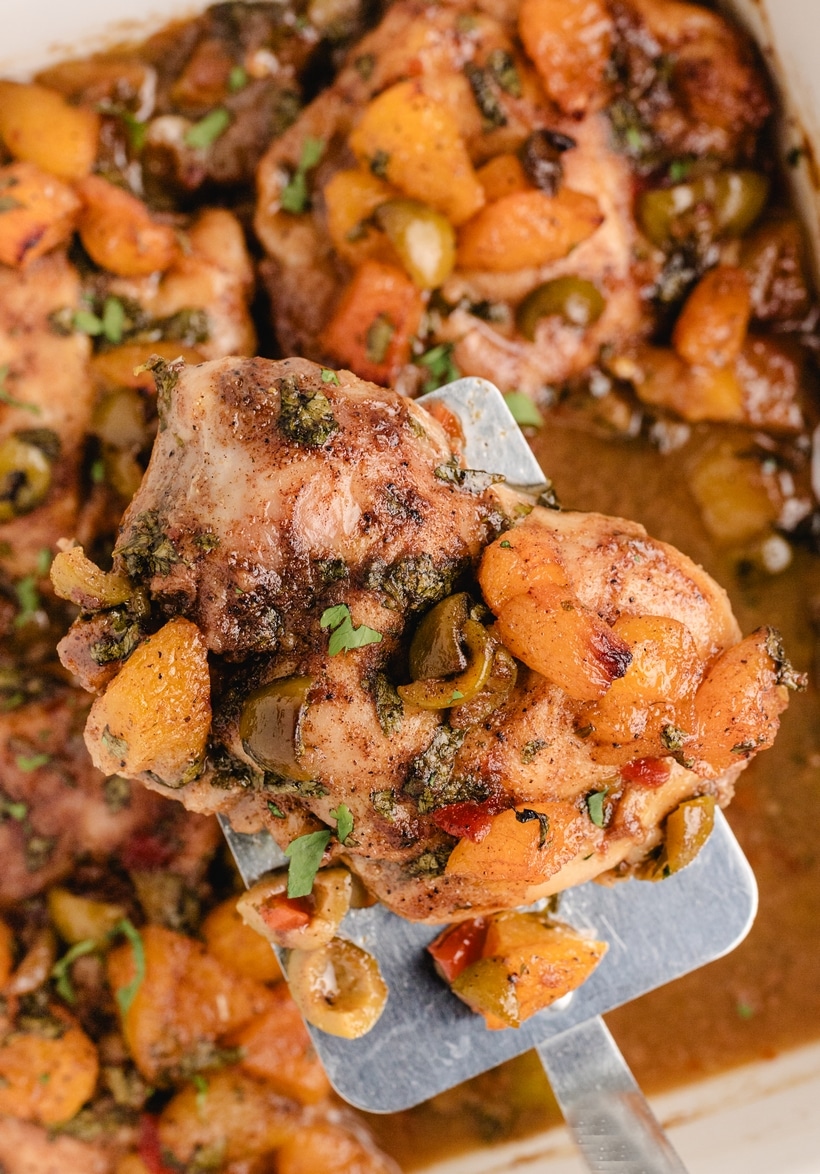 chicken thigh with apricots and olives on spatula Baked chicken thigh lifted from a pan, topped with dried apricots, olives, and herbs.