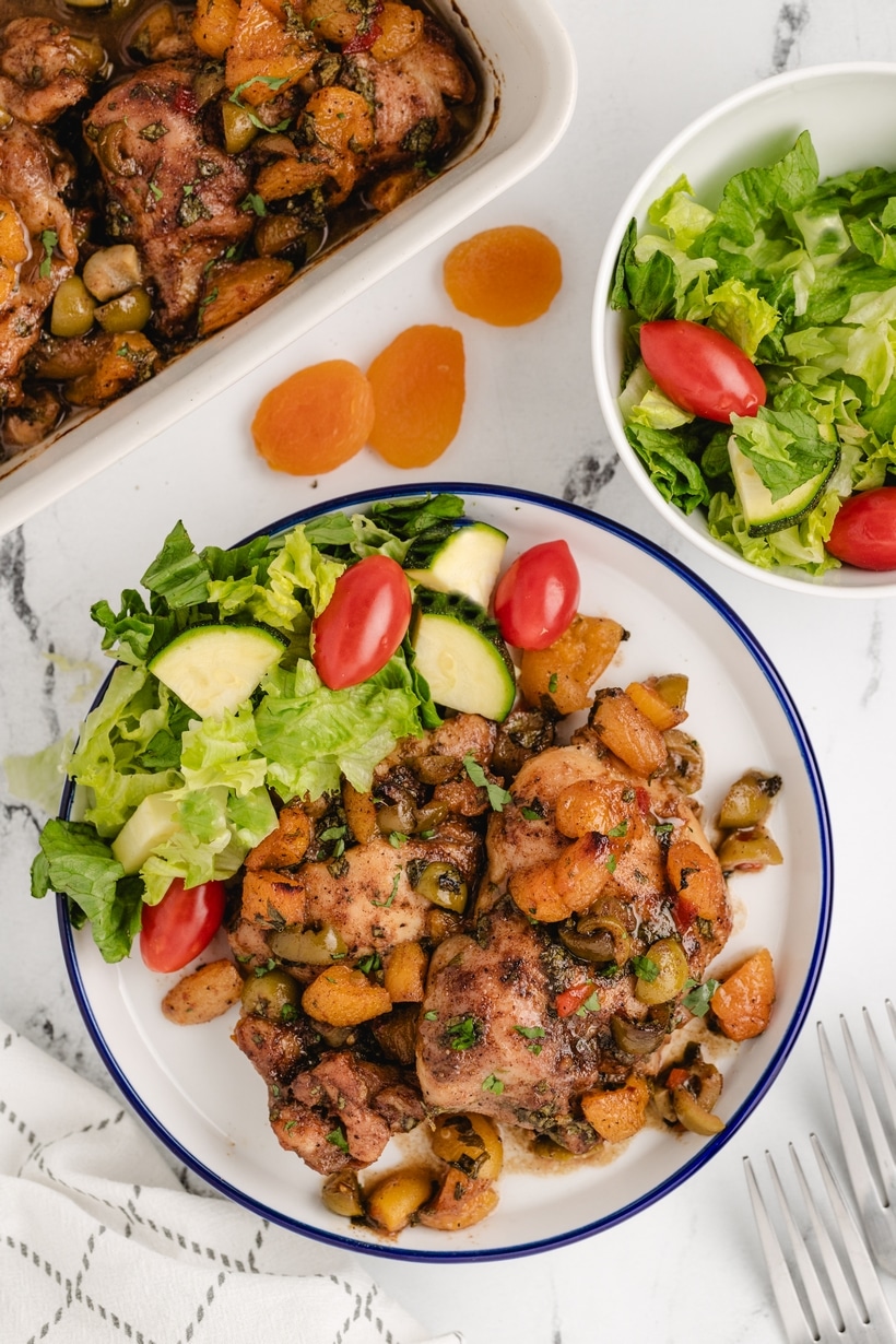 apricot chicken thighs with salad and couscous on plate Two apricot chicken thighs served with couscous and side salad of romaine, tomatoes, and cucumbers.