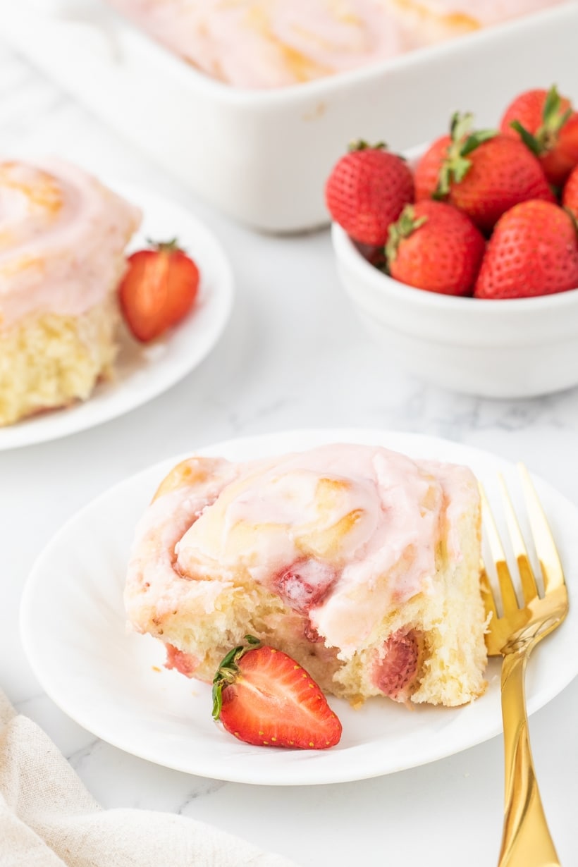 A single strawberry sweet roll on a white plate, garnished with fresh strawberries on the side.