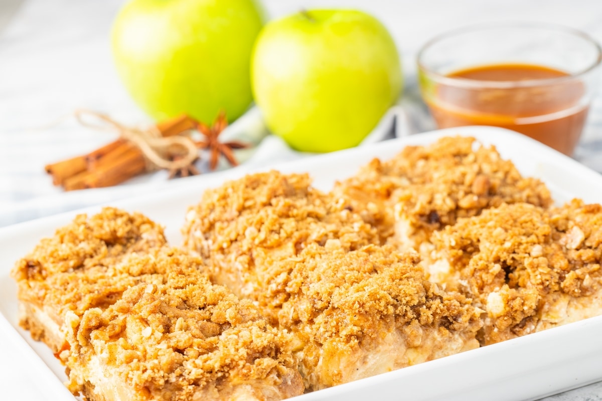 Apple cheesecake bars in baking dish. Apple cheesecake bars with streusel topping lined up in a white baking dish.