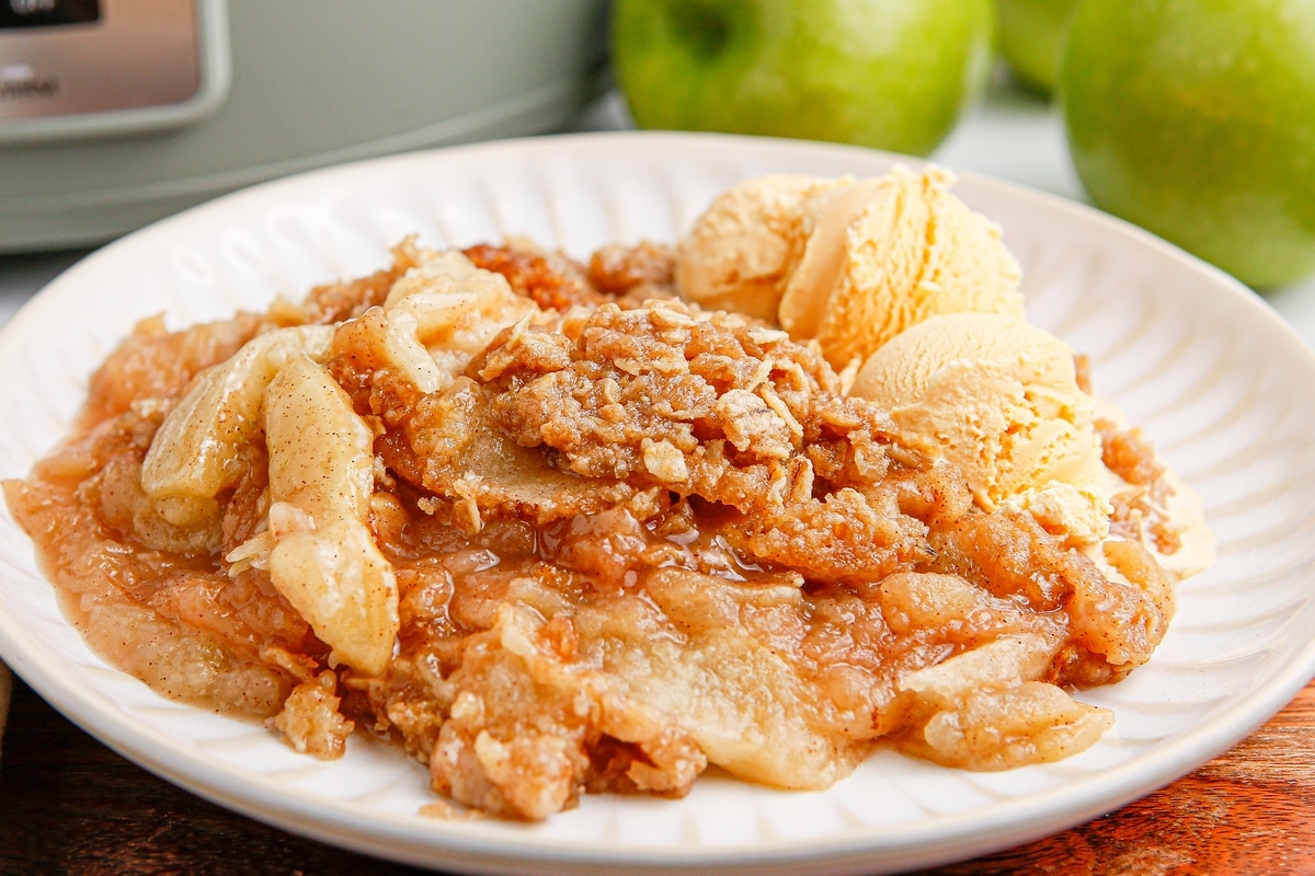 Slow cooker apple crisp served with ice cream on a plate in front of the crockpot and fresh green apples.