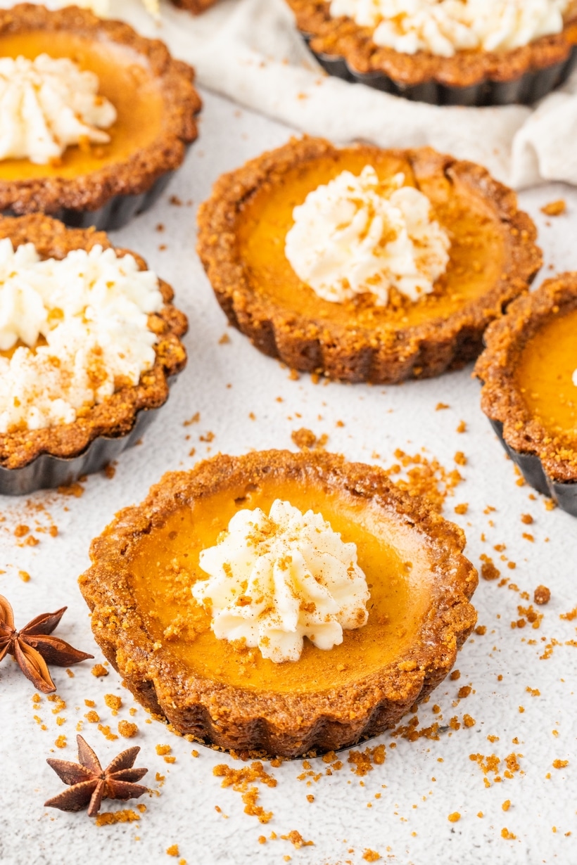 Mini pumpkin tarts with gingersnap crust and whipped cream topping, dusted with cookie crumbs on a white surface.