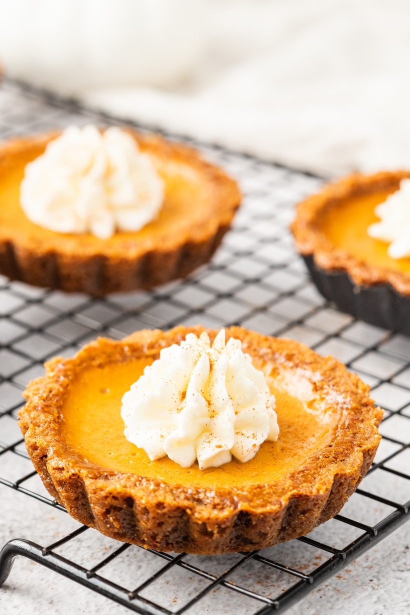 Mini pumpkin tarts cooling on a wire rack after baking, each topped with whipped cream and sprinkled with spice.