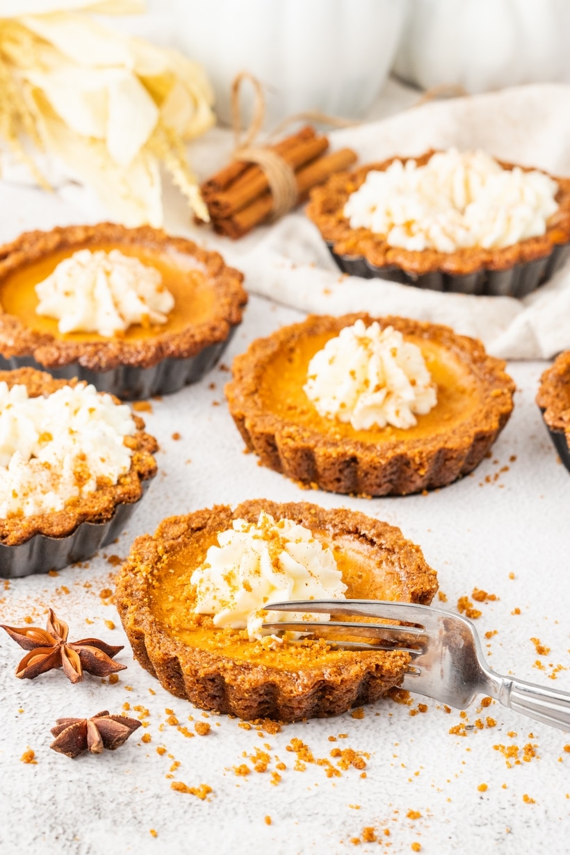 Mini pumpkin tarts with gingersnap crust and whipped cream, sprinkled with crumbs and cinnamon, served with a fork on a white surface.