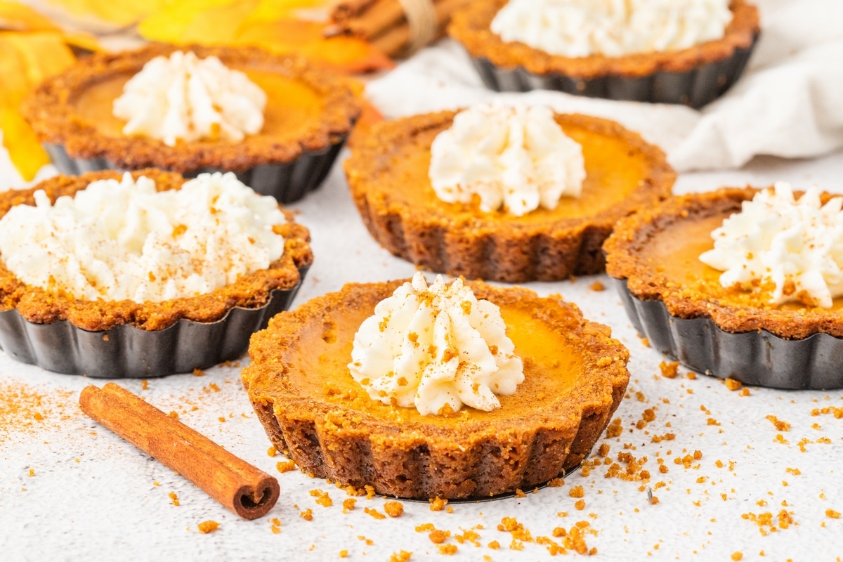 Mini pumpkin tarts with gingersnap crust and whipped cream, surrounded by cinnamon and fall leaves on a light surface.