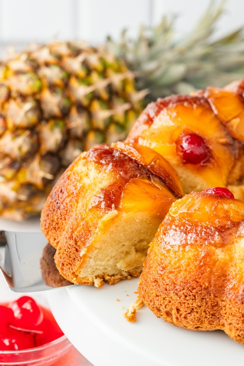 Serving Pineapple Upside Down Cake from a Bundt Pan Pineapple upside down Bundt cake being lifted from a cake stand, with a whole pineapple in the background.