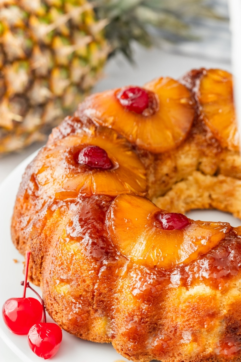 Pineapple Upside Down Bundt Cake with Caramelized Topping Side view of pineapple upside down cake with glossy pineapple and cherry topping, whole pineapple in background.