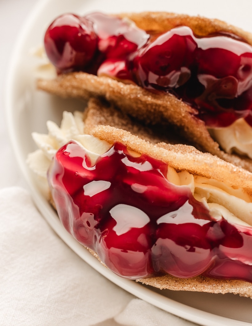 Top view of two cherry cheesecake tacos on a white plate, filled with cheesecake and topped with cherry pie filling.