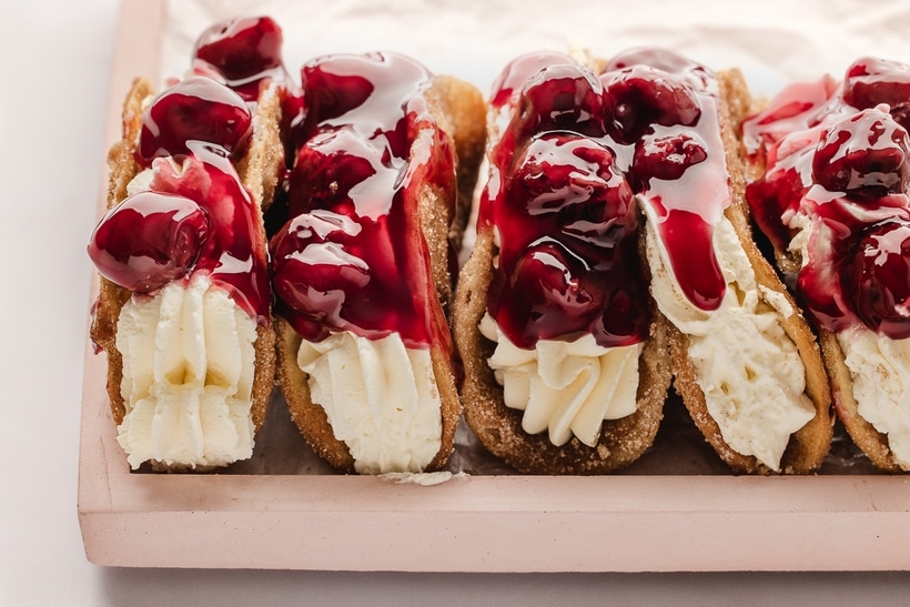 Full view of multiple cherry cheesecake tacos standing upright, filled with cheesecake and topped with cherry pie filling.