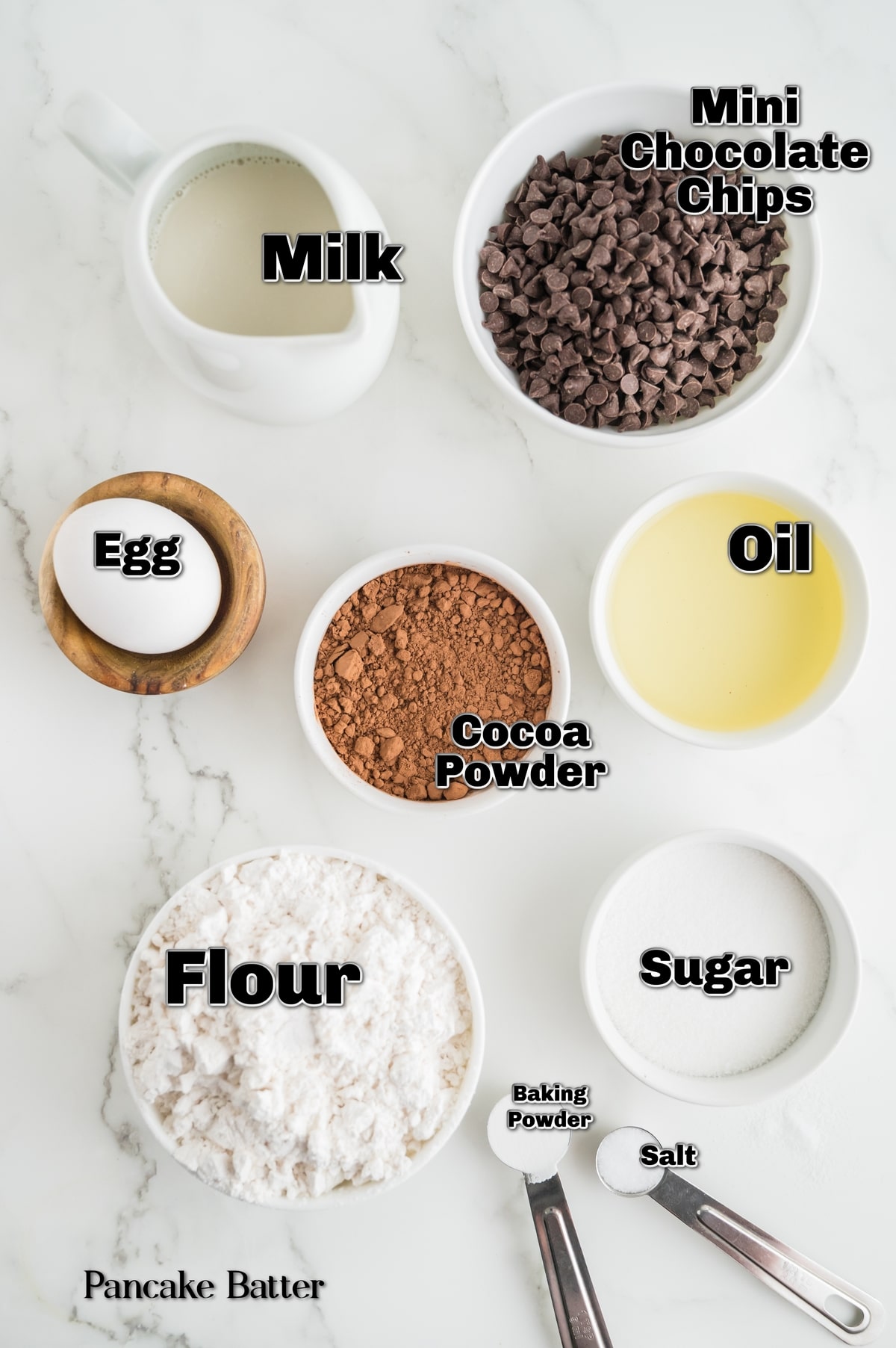 Flat lay of measured ingredients for chocolate pancake batter arranged on a marble surface, including flour, cocoa powder, milk, egg, vegetable oil, sugar, baking powder, salt, and mini chocolate chips. Ingredients for double chocolate pancake batter including flour, cocoa powder, milk, egg, oil, sugar, baking powder, salt, and mini chocolate chips