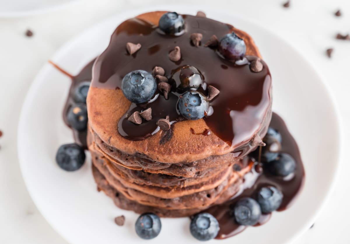 Top-down view of a stack of chocolate pancakes covered in warm chocolate ganache and finished with blueberries and mini chocolate chips on a white plate. Overhead view of chocolate pancakes topped with warm chocolate ganache, blueberries, and mini chocolate chips
