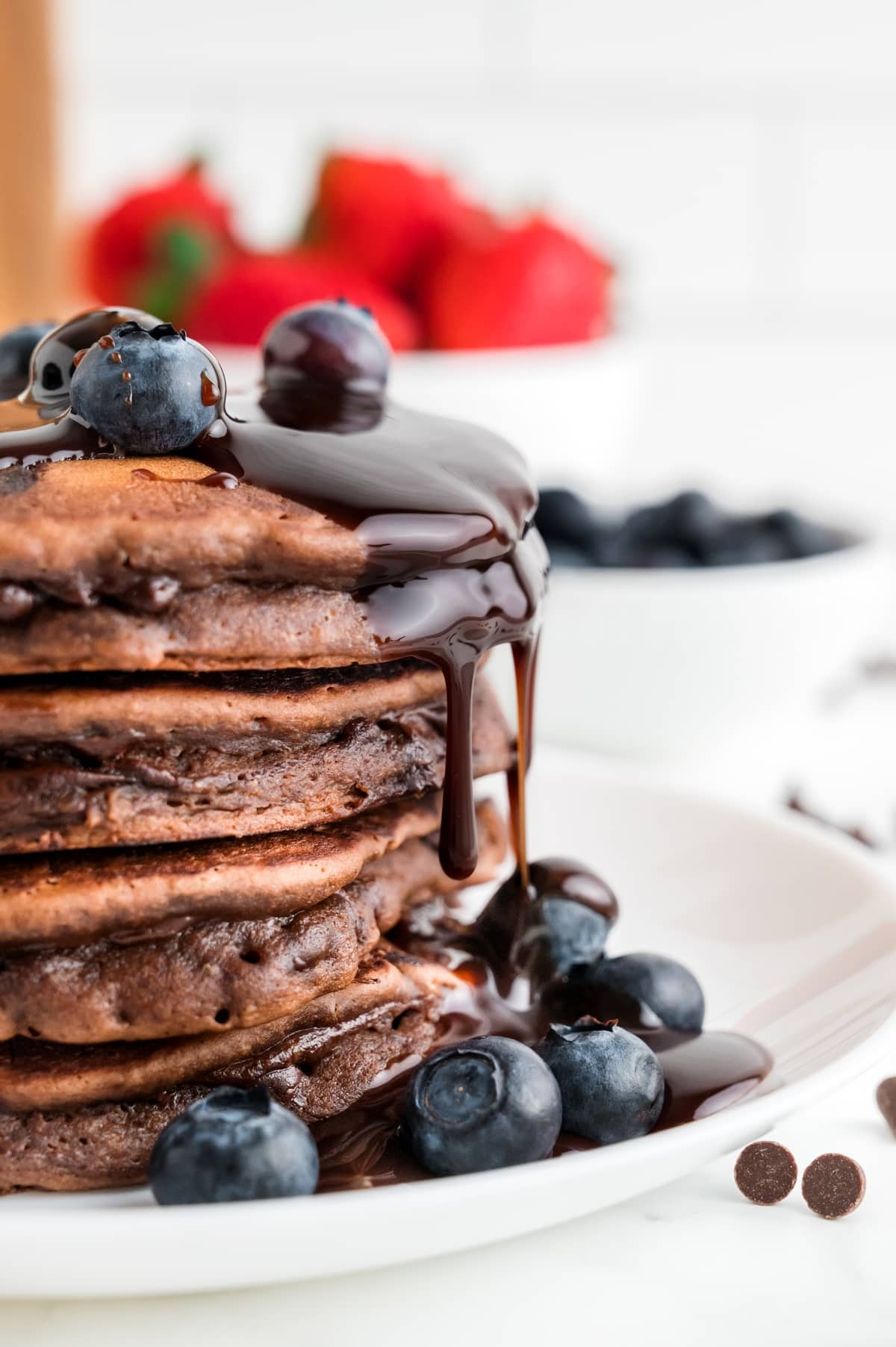 A tall stack of homemade chocolate pancakes topped with rich chocolate ganache dripping down the sides and finished with fresh blueberries on a white plate. Stack of chocolate pancakes with glossy chocolate ganache and fresh blueberries on a white plate