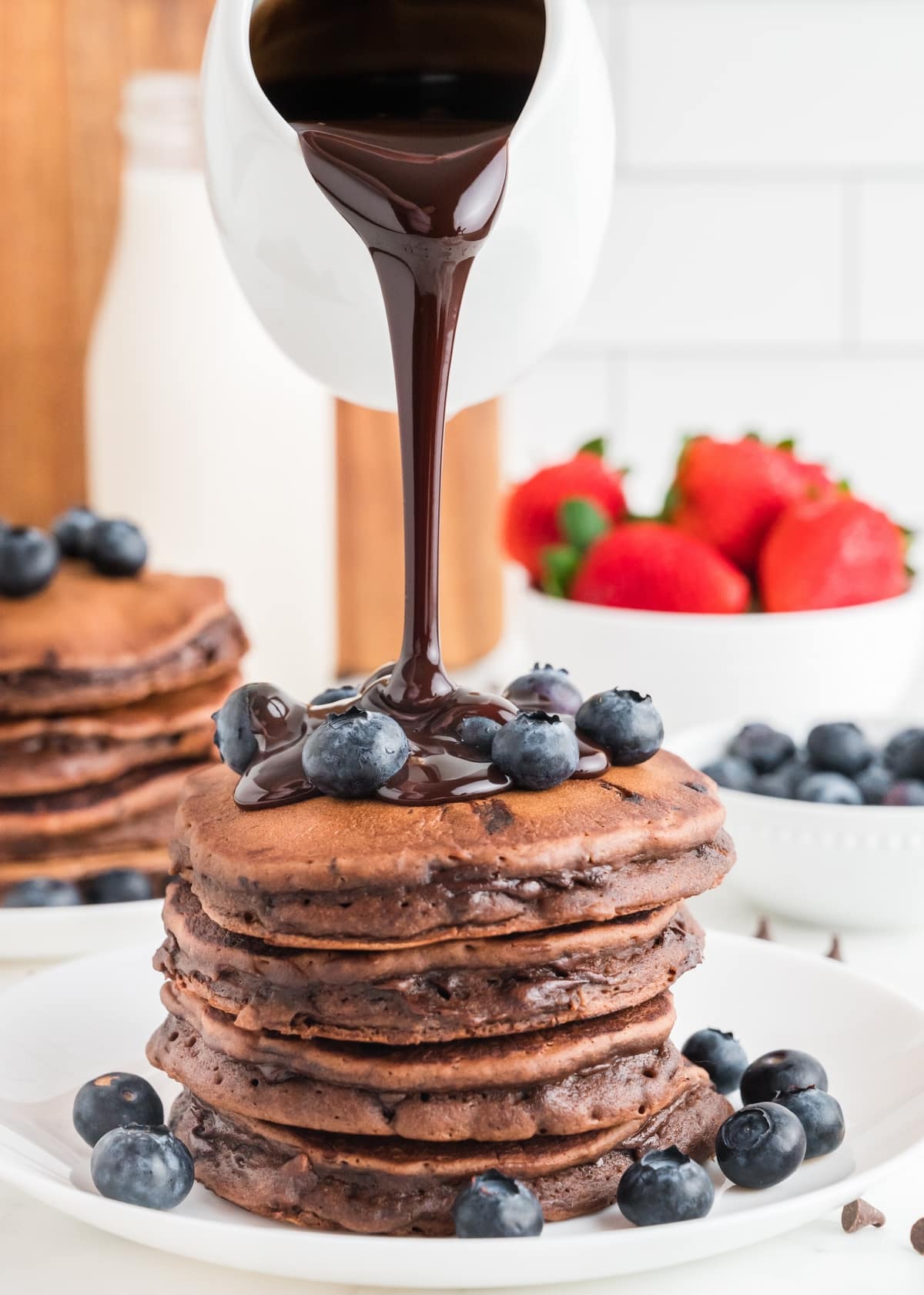 Warm ganache pouring over a tall stack of chocolate pancakes. Chocolate pancakes stacked high with warm chocolate ganache being poured over the top and fresh blueberries