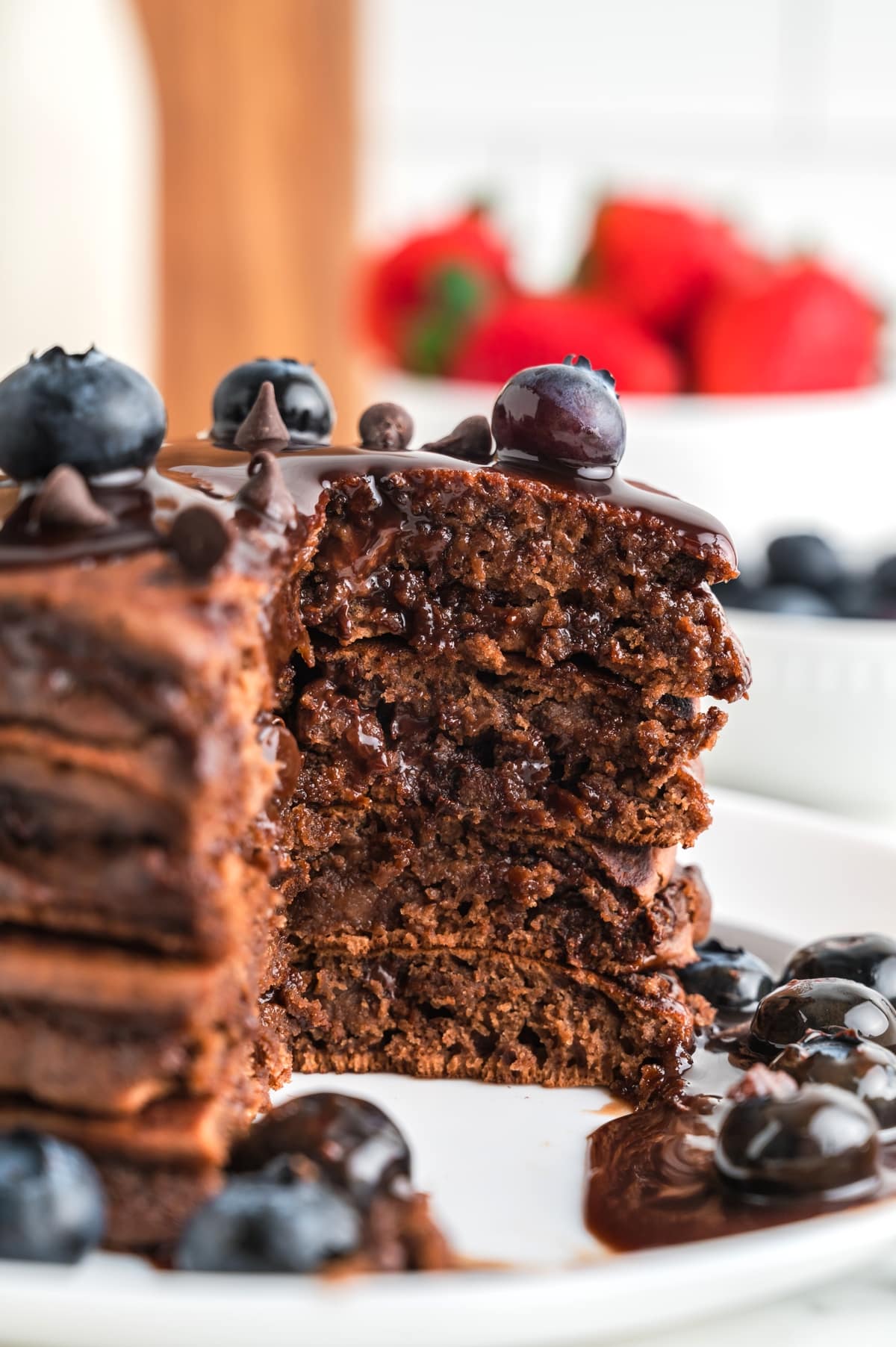 Tight close-up of sliced chocolate pancakes showing a moist interior filled with melted chocolate chips, topped with glossy ganache and blueberries on a white plate. Close-up of chocolate pancakes with melted chocolate chips inside, topped with ganache and fresh blueberries