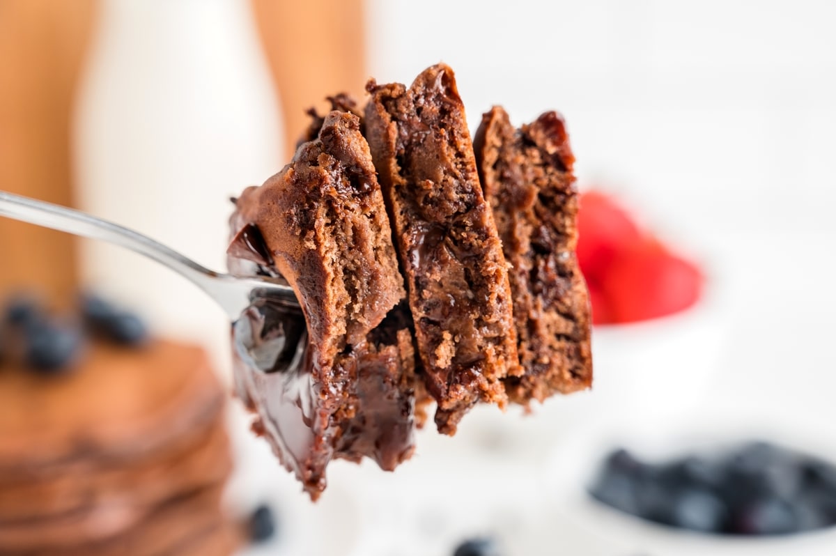 Close-up of a fork lifting a thick slice of double chocolate pancake showing melted chocolate chips inside and coated in warm ganache, with fresh berries blurred in the background. Fork holding a thick bite of double chocolate pancake with melted chocolate and ganache