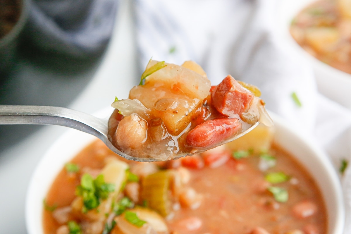 Close-up of a spoon lifting soup above the bowl, showing the texture of beans, ham, and veggies.