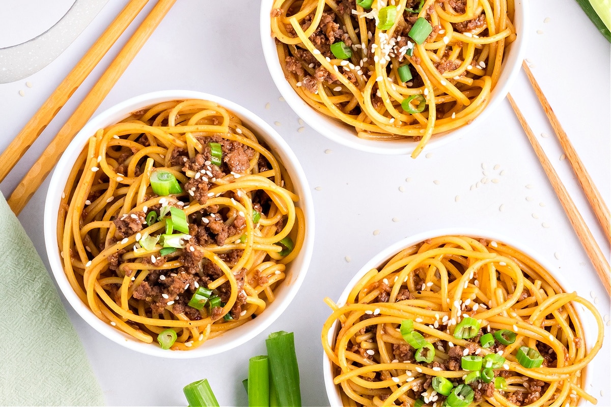 Three bowls of Mongolian beef noodles garnished with sesame seeds and green onions on a white background by Noble Pig.