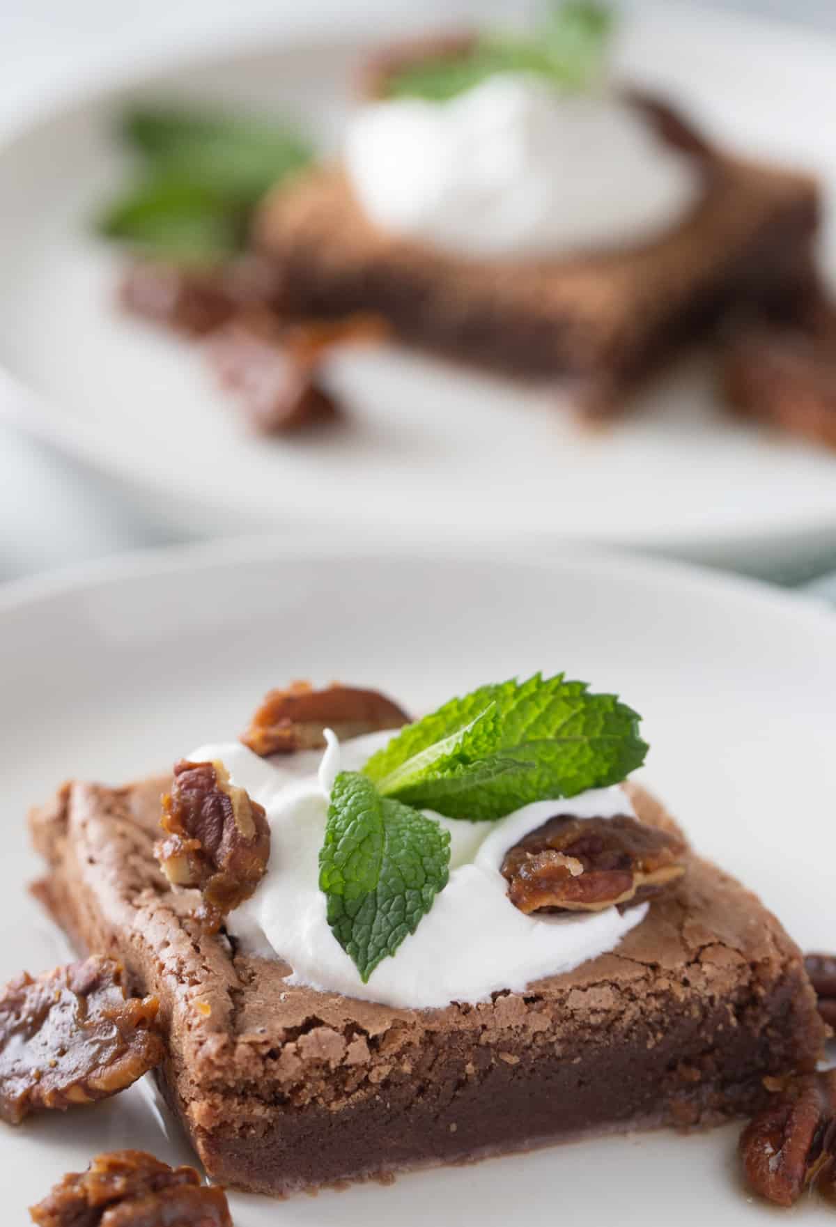 Brownie on a plate topped with whipped cream, candied pecans, and mint, with another dressed brownie blurred in the back