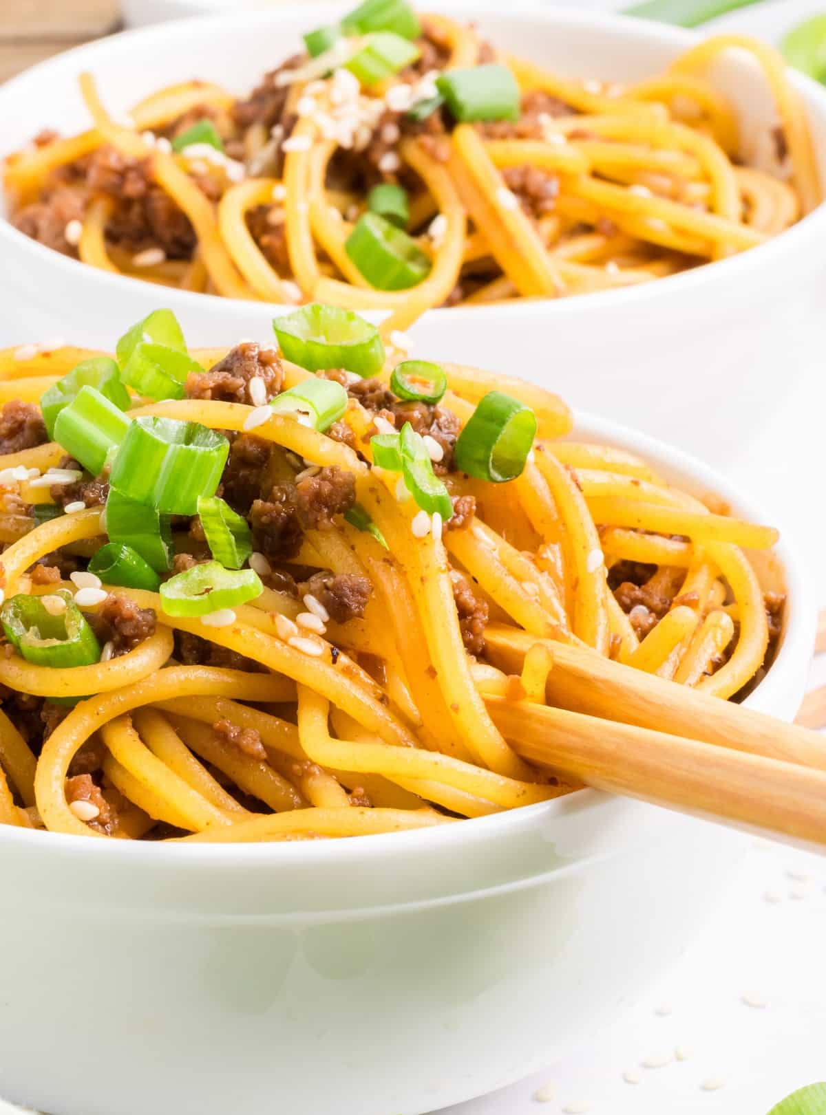 Bowl of Mongolian beef noodles topped with green onions and sesame seeds, sauce-coated noodles twirled with chopsticks by Noble Pig.